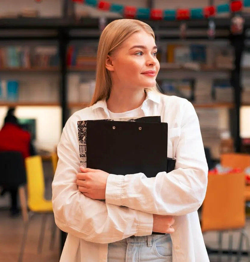 młoda kobieta z czarnym notesem spogląda w bok. W tle regały z ksiażkami i biurkami